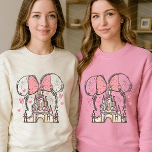 Pu&ograve; includere: Due donne indossano felpe color crema e rosa con una grafica di un castello, un fiocco e palloncini. La grafica &egrave; un disegno stilizzato di un castello con un fiocco e palloncini su entrambi i lati.