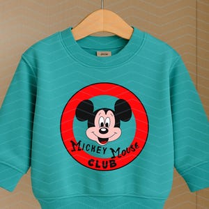 May include: A teal long-sleeve sweatshirt featuring a subtle chevron pattern and a central graphic of Mickey Mouse inside a red circle. The text "MICKEY MOUSE CLUB" is printed in black lettering below the character's face on the front of the garment.