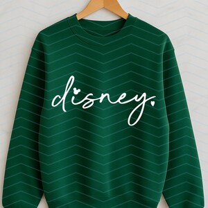 Op de afbeelding: Een donkergroene sweatshirt met een wit cursief "disney." tekstontwerp. Het woord "disney" heeft een kleine Mickey Mouse-kop die de "i" vervangt. De sweatshirt hangt aan een houten kleerhanger.