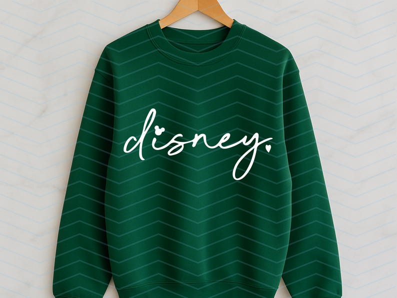 Op de afbeelding: Een donkergroene sweatshirt met een wit cursief "disney." tekstontwerp. Het woord "disney" heeft een kleine Mickey Mouse-kop die de "i" vervangt. De sweatshirt hangt aan een houten kleerhanger.