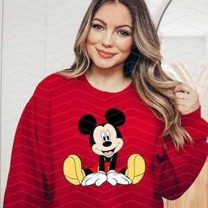May include: A red sweatshirt featuring a cartoon image of Mickey Mouse in the centre. Mickey Mouse is wearing red shorts and yellow shoes. He is smiling and has large, black ears.