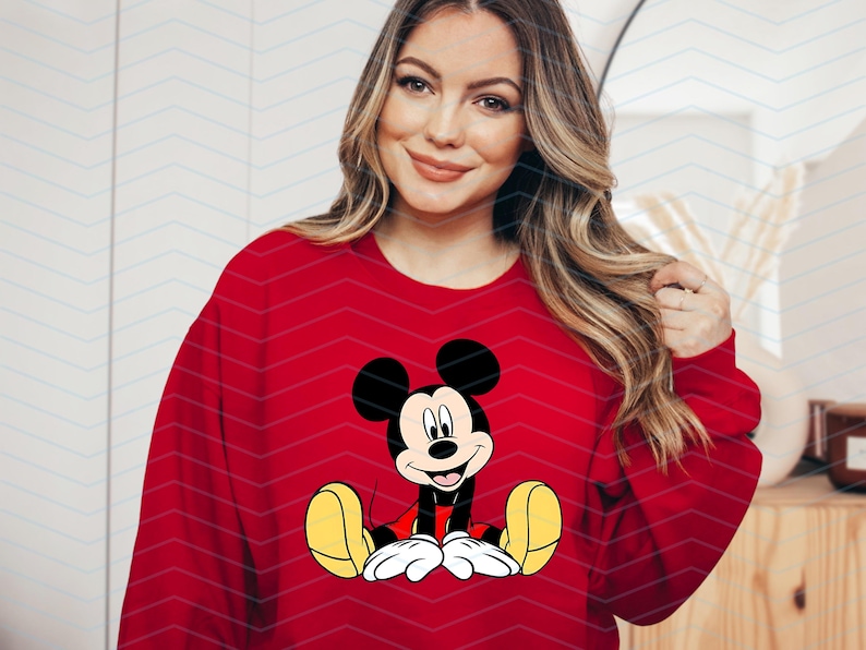 May include: A red sweatshirt featuring a cartoon image of Mickey Mouse in the centre. Mickey Mouse is wearing red shorts and yellow shoes. He is smiling and has large, black ears.