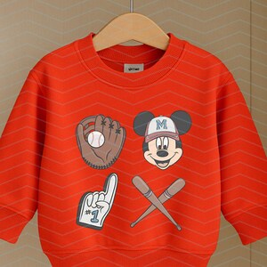 May include: Red sweatshirt with a Mickey Mouse graphic wearing a baseball cap with the letter "M" on it. The graphic also includes a baseball, a baseball glove, and two crossed baseball bats. The sweatshirt has a crew neck and long sleeves.