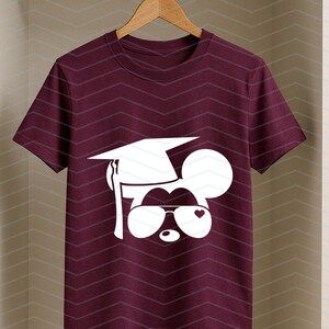 May include: Burgundy t-shirt featuring a white graphic of a mouse wearing a graduation cap and sunglasses with a heart. The shirt is hanging on a wooden hanger against a chevron patterned background.