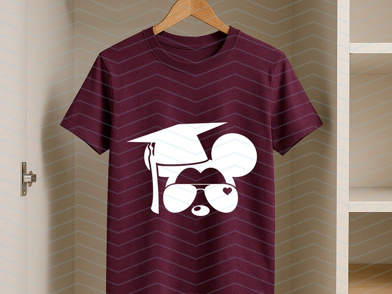 May include: Burgundy t-shirt featuring a white graphic of a mouse wearing a graduation cap and sunglasses with a heart. The shirt is hanging on a wooden hanger against a chevron patterned background.