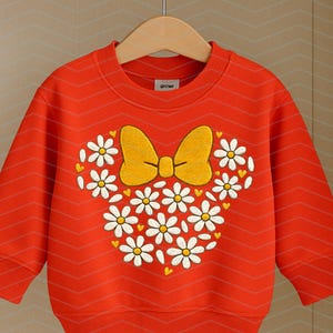 Puede incluir: Una sudadera roja brillante con un dise&ntilde;o en forma de margarita, un lazo amarillo y margaritas blancas. La sudadera tiene mangas largas y cuello redondo. El dise&ntilde;o est&aacute; centrado en la parte delantera.
