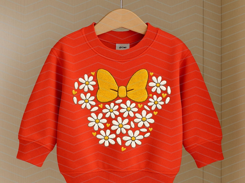 Puede incluir: Una sudadera roja brillante con un dise&ntilde;o en forma de margarita, un lazo amarillo y margaritas blancas. La sudadera tiene mangas largas y cuello redondo. El dise&ntilde;o est&aacute; centrado en la parte delantera.