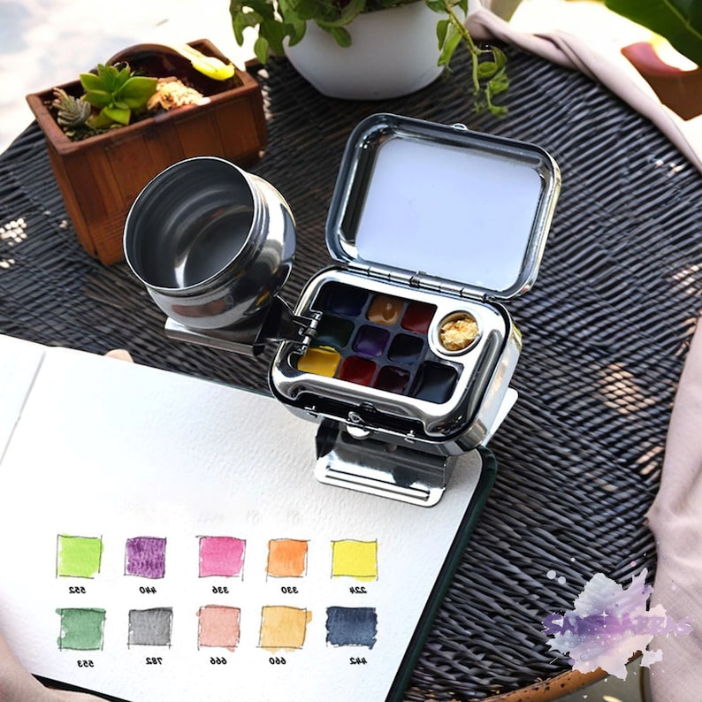 May include: A silver metal watercolor paint set with a built-in water container. The set has 12 paint pans and a separate well for a brush. The set is open and sitting on a white surface.
