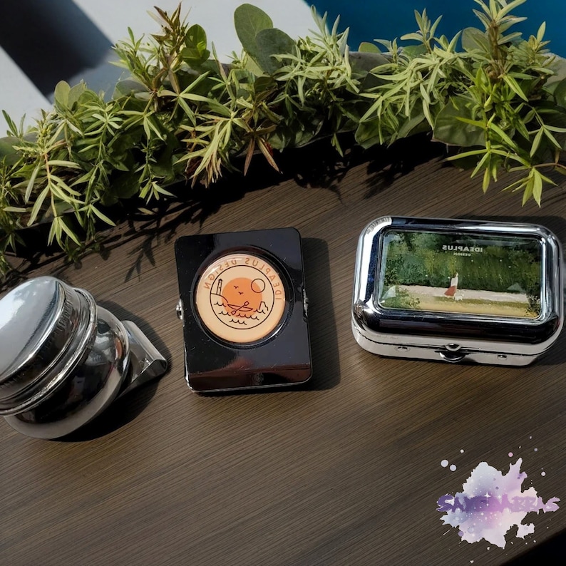 May include: Three small, silver, rectangular boxes. The box on the left has a black lid with a round, orange and brown sticker that says "Holiday Sunset". The box in the middle is empty. The box on the right has a clear lid with a picture of a woman walking in a green forest. The boxes are on a wooden surface.