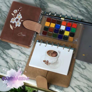 May include: A brown leather bound journal with a floral design and the word "Cash" written on the cover. The journal is open to a page with a watercolor painting of a cup of coffee. A watercolor paint set is visible in the background.