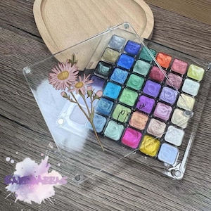 Könnte beinhalten: Eine transparente Acrylpalette mit einem floralen Design und dem Wort "Chance" darauf. Die Palette hat 36 kleine Quadrate, die mit bunten Metallic-Farben gefüllt sind.