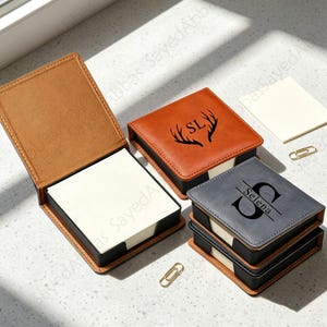 Könnte beinhalten: Drei quadratische Notizzettelhalter mit Papierstapeln. Ein beigefarbener Halter ist geöffnet, ein beigefarbener ist mit einem Geweihmotiv und den Buchstaben SL geschlossen, und ein grauer ist mit einem großen S und dem Namen Selena bedruckt.