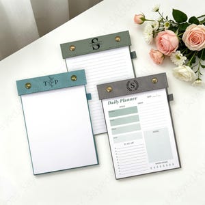 Könnte beinhalten: Drei personalisierte Notizblockhalter mit lederartigen Bezügen in Türkis, Grau und Grün. Jeder Notizblock hat ein anderes Design, darunter liniertes Papier, ein Tagesplaner-Layout und eine leere Seite. Die Umschläge sind mit Initialen und dekorativen Akzenten verziert.