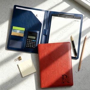 May include: A navy blue leather-like portfolio folder containing a calculator, card slots, and a lined memo pad, paired with a closed rust-colored notebook featuring the letter R and the name Roberta, a gold paper clip, a sticky note, and two pens.