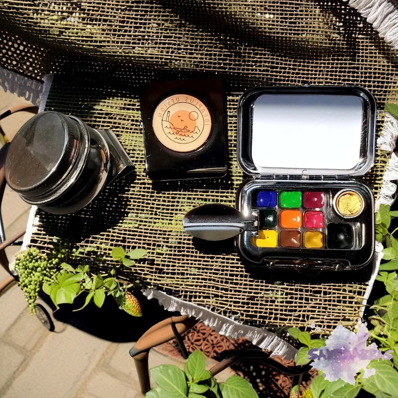 May include: A silver metal watercolor paint set with a mirror lid and a small round container of gold paint. The set is open and shows the 9 watercolor pans. The set is on a brown woven surface with a small jar and a round sticker with the text "TOGETHER DESIGN" on it.