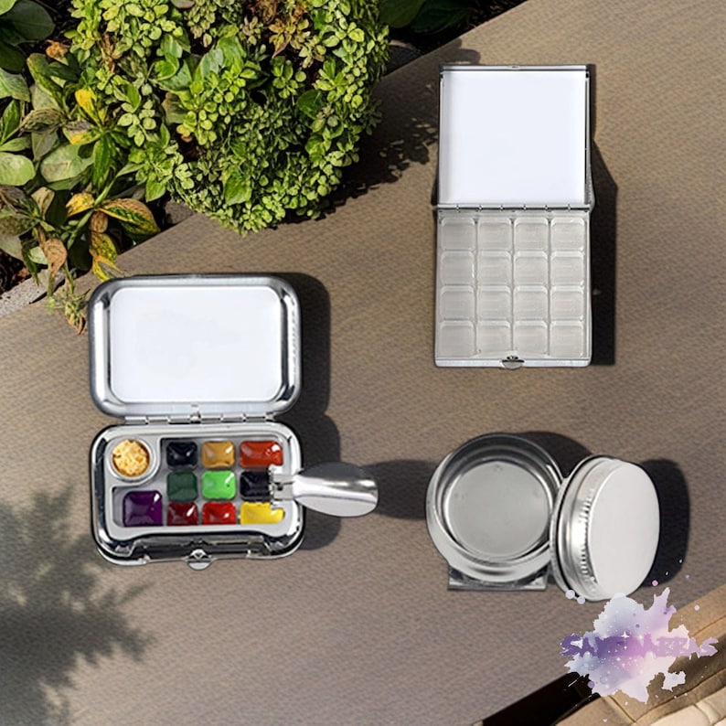 May include: A silver metal watercolor paint palette with a hinged lid and a brush. The palette has 12 paint wells filled with colorful watercolor paints. The palette is open and the lid is propped up. There is a small silver metal container with a lid and a silver metal brush.