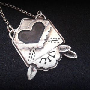 Puede incluir: Un collar de plata con un colgante en forma de sobre con un coraz&oacute;n en su interior. El sobre tiene un borde festoneado y est&aacute; decorado con un dise&ntilde;o floral. El coraz&oacute;n es negro y tiene un borde festoneado.