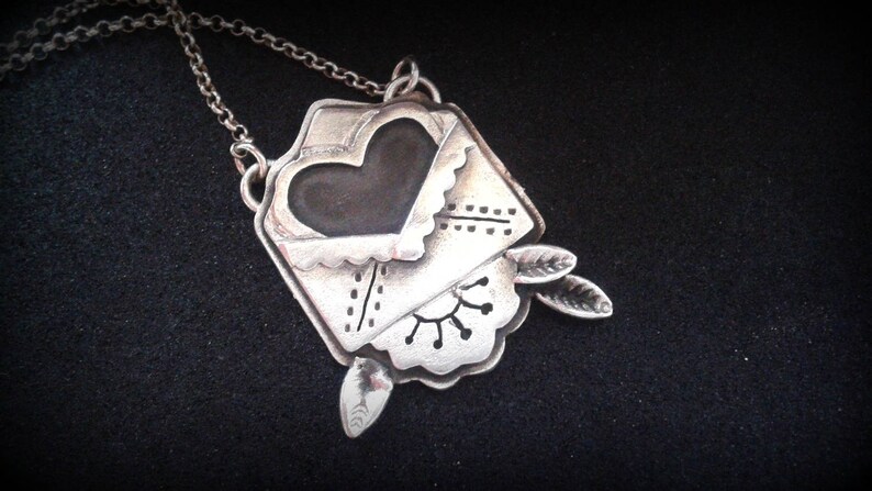 Puede incluir: Un collar de plata con un colgante en forma de sobre con un coraz&oacute;n en su interior. El sobre tiene un borde festoneado y est&aacute; decorado con un dise&ntilde;o floral. El coraz&oacute;n es negro y tiene un borde festoneado.