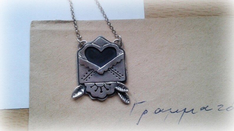 Puede incluir: Collar de plata con un colgante en forma de coraz&oacute;n que parece un sobre. El sobre tiene un dise&ntilde;o decorativo y est&aacute; abierto para revelar el coraz&oacute;n.