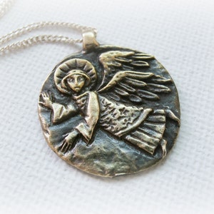 May include: A silver chain necklace with a round bronze pendant featuring a detailed relief sculpture of an angel with wings outstretched.