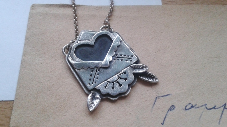 Puede incluir: Un collar de plata con un colgante en forma de sobre con un coraz&oacute;n en su interior. El sobre tiene un dise&ntilde;o detallado y el coraz&oacute;n es negro.