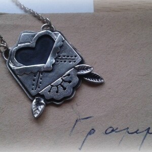 Puede incluir: Un collar de plata con un colgante en forma de sobre con un coraz&oacute;n en su interior. El sobre tiene un dise&ntilde;o decorativo y el coraz&oacute;n es negro.