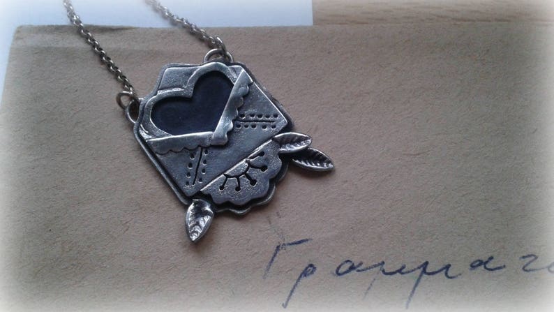 Puede incluir: Un collar de plata con un colgante en forma de sobre con un coraz&oacute;n en su interior. El sobre tiene un dise&ntilde;o decorativo y el coraz&oacute;n es negro.