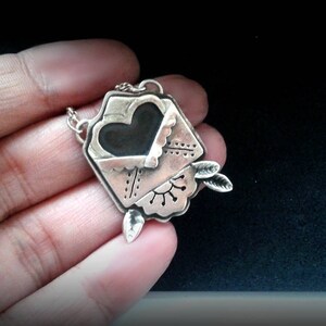 Puede incluir: Un collar de plata con un colgante en forma de sobre con un coraz&oacute;n en su interior. El sobre tiene un dise&ntilde;o detallado y el coraz&oacute;n es negro.