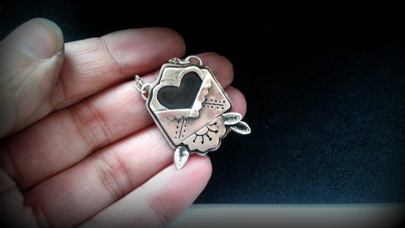 Puede incluir: Un collar de plata con un colgante en forma de sobre con un coraz&oacute;n en su interior. El sobre tiene un dise&ntilde;o detallado y el coraz&oacute;n es negro.