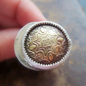 May include: A silver ring with a gold-toned circular design featuring a starburst pattern and intricate details.