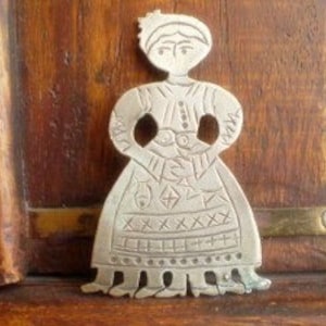 May include: A silver charm in the shape of a woman wearing a long dress with a geometric pattern. The charm has a simple, folk art style.