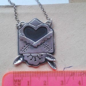 Puede incluir: Collar de plata con un colgante en forma de coraz&oacute;n que parece un sobre. El colgante tiene un dise&ntilde;o detallado con un coraz&oacute;n en el centro y hojas en la parte inferior.