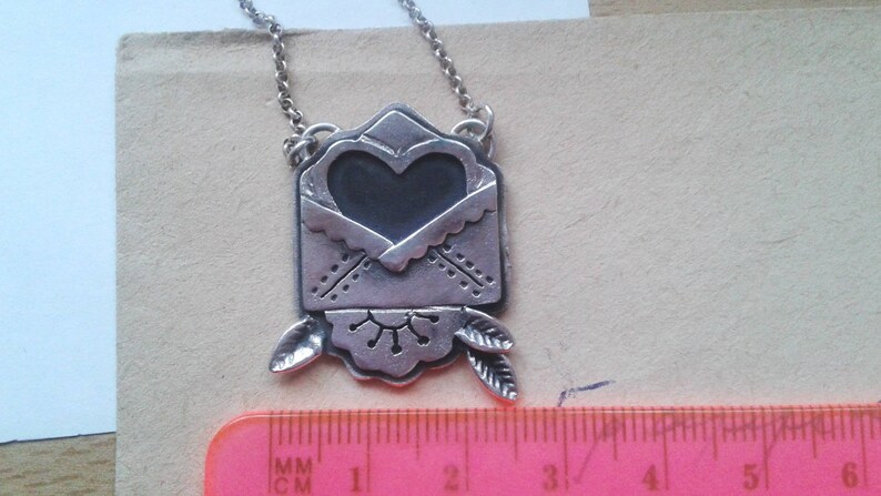 Puede incluir: Collar de plata con un colgante en forma de coraz&oacute;n que parece un sobre. El colgante tiene un dise&ntilde;o detallado con un coraz&oacute;n en el centro y hojas en la parte inferior.