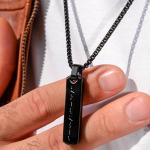 Puede incluir: Un collar de cadena negra con un colgante negro que presenta una línea vertical de puntos y rayas en código Morse.