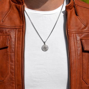 Może przedstawiać: Srebrny łańcuszek z kompasem jako zawieszką. Łańcuszek nosi osoba w brązowej sk&oacute;rzanej kurtce i białym t-shircie.