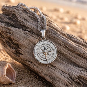 May include: A silver-toned metal pendant necklace featuring a circular compass design with cardinal directions N, E, S, and W engraved on the face. The pendant hangs from a matching silver-toned chain link necklace with a polished metallic finish.