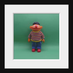 May include: A plush toy of a Sesame Street character, Bert, wearing a striped jumper and blue jeans. He has a brown face, black hair, and a red nose.