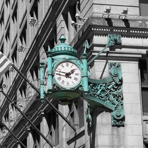 May include: A teal-colored clock face with black Roman numerals and hands, mounted on a building with a black and white facade. The clock is part of a decorative, ornate, teal-colored metal structure.