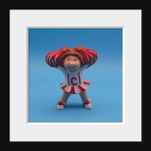 May include: A vintage Cabbage Patch Kids cheerleader figurine. The doll has red hair and is wearing a white and red cheerleader uniform with the letter "C". The figurine is set against a blue background.