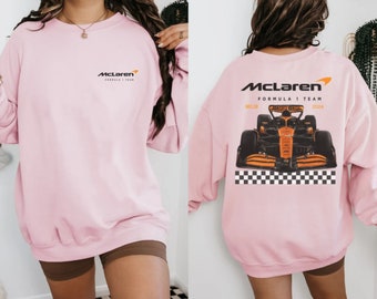May include: A light pink sweatshirt featuring the McLaren Formula 1 Team logo on the front and a graphic of a McLaren race car on the back. The back design includes the text "McLaren Formula 1 Team" and "2024".