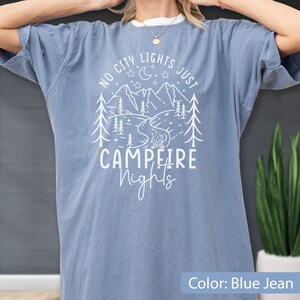 May include: A blue jean coloured short-sleeve t-shirt featuring a white graphic print of mountains, pine trees, a crescent moon, stars, and a campfire. The text reads NO CITY LIGHTS JUST CAMPFIRE NIGHTS in a mix of block and script lettering.