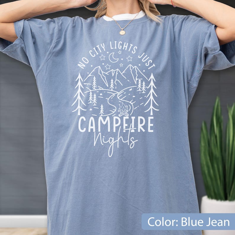 May include: A blue jean coloured short-sleeve t-shirt featuring a white graphic print of mountains, pine trees, a crescent moon, stars, and a campfire. The text reads NO CITY LIGHTS JUST CAMPFIRE NIGHTS in a mix of block and script lettering.