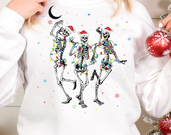May include: A white long-sleeve sweatshirt featuring a graphic of three dancing skeletons wearing red Santa hats. The skeletons are wrapped in colorful Christmas light strings. The design includes small red, blue, and green snowflakes and stars.