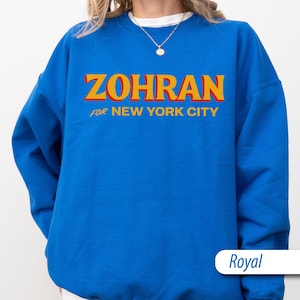 May include: Royal blue sweatshirt with the word "ZOHRAN" in large, yellow, and red letters. Below the word "ZOHRAN" is the text "FOR NEW YORK CITY". The sweatshirt is a solid color and has a crew neck.