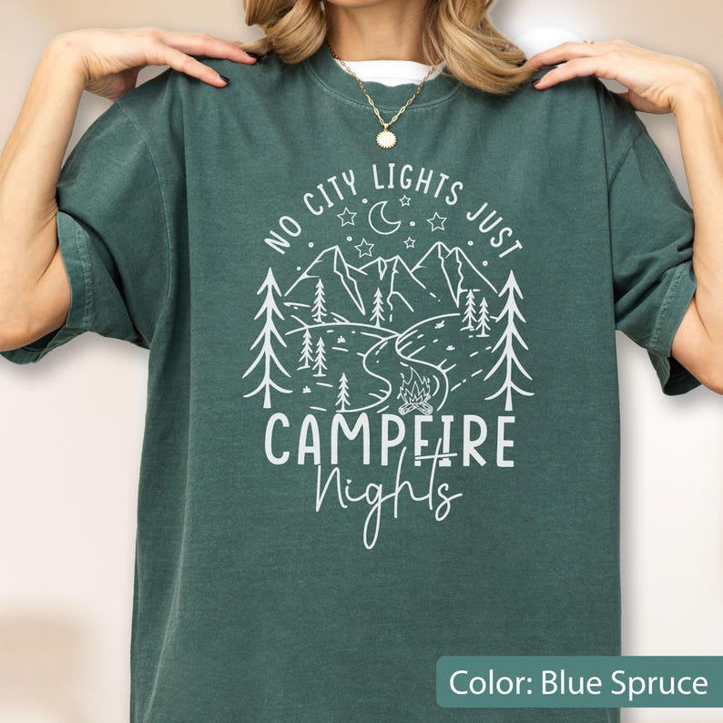 May include: A blue spruce short-sleeve t-shirt featuring a white graphic print of mountains, pine trees, a crescent moon, stars, and a campfire. The text reads NO CITY LIGHTS JUST CAMPFIRE Nights in a mix of block and script lettering.