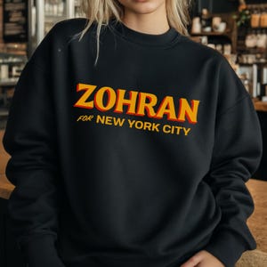 May include: Black crew neck sweatshirt with the text "ZOHRAN" in yellow and red, with "FOR NEW YORK CITY" below. The sweatshirt is a solid black color.