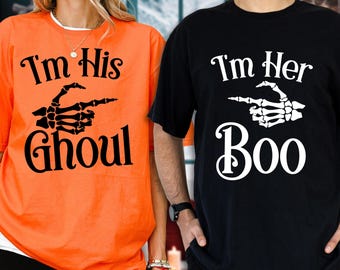 May include: An orange t-shirt with black text reading I'm His Ghoul and a black skeleton hand graphic, and a black t-shirt with white text reading I'm Her Boo and a white skeleton hand graphic, both featuring short sleeves and crew necklines.