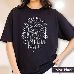 May include: A black short-sleeved t-shirt featuring a white graphic print of mountains, pine trees, a crescent moon, stars, and a campfire. The text reads NO CITY LIGHTS JUST CAMPFIRE Nights in a mix of block and script lettering on the front.