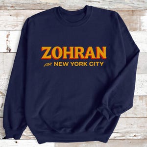 May include: Navy blue sweatshirt with the text "ZOHRAN" in orange and yellow, with the words "FOR NEW YORK CITY" in yellow underneath. The sweatshirt is a crew neck style.