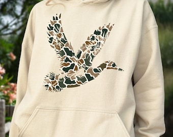 May include: A cream-colored hooded sweatshirt featuring a large graphic of a flying duck on the chest. The duck silhouette is filled with a camouflage pattern consisting of various shades of brown, dark green, and tan shapes.